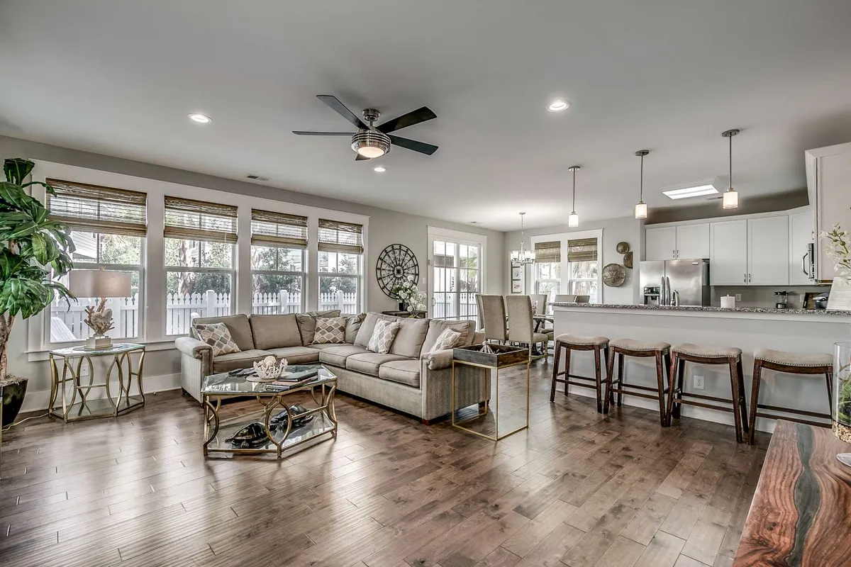 A comfortable open-plan living space featuring a plush sofa, coffee table, bar stools, and a visible kitchen area. Large windows fill the room with natural light, while the layout encourages flow between relaxing, dining, and cooking spaces.