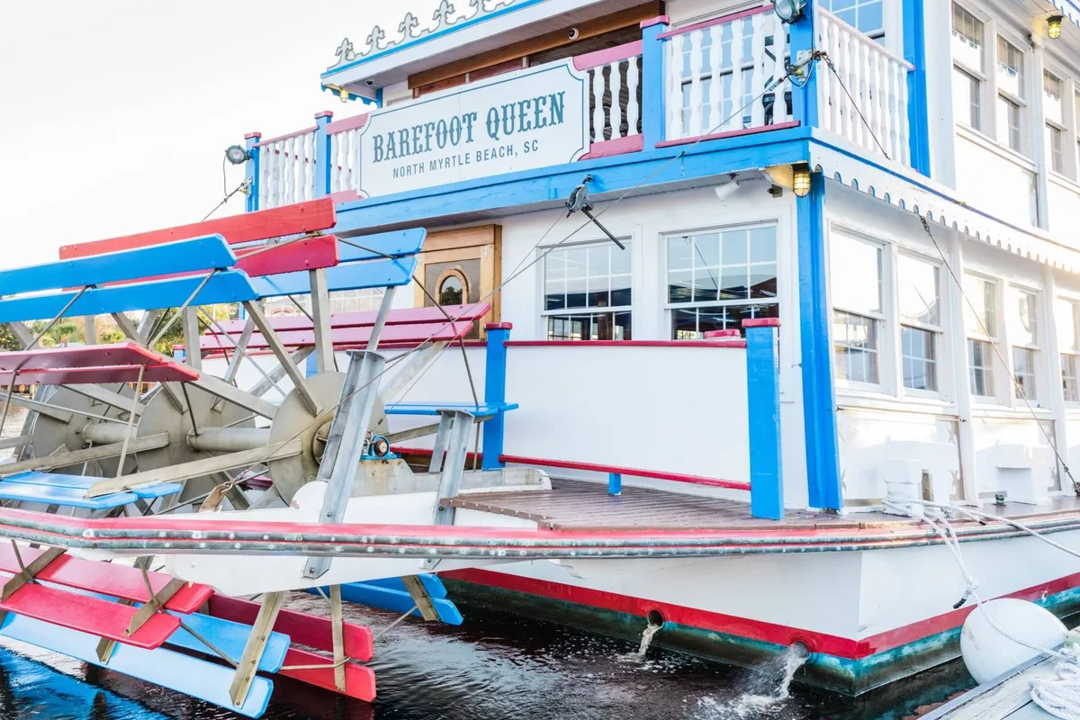 A colorful and inviting exterior shot of the Barefoot Queen riverboat, with its distinctive paddle wheel and vibrant signage. The lively mood suggests opportunities for waterfront dining, sightseeing, and evening entertainment.