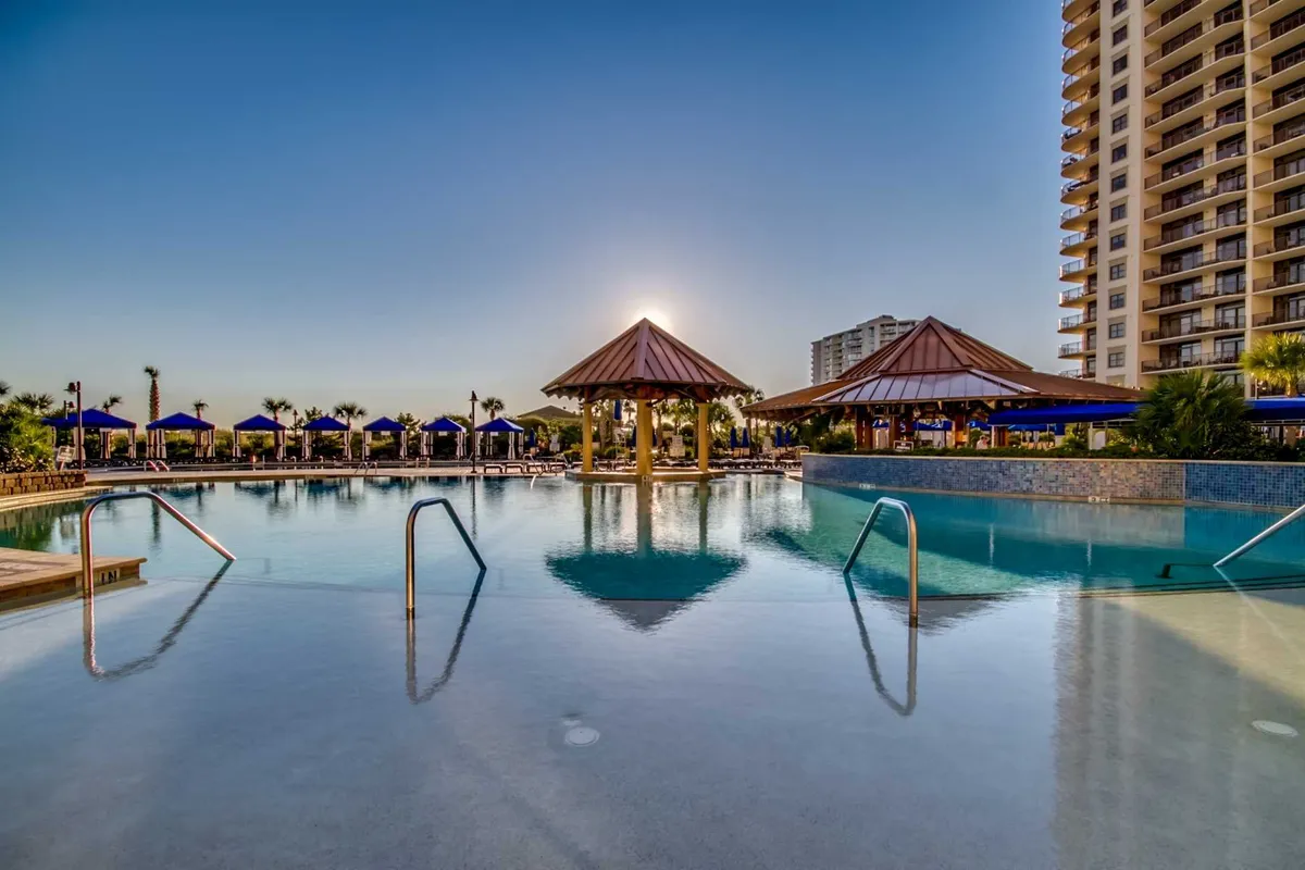 This image showcases an inviting outdoor swimming pool surrounded by thatched-roof cabanas with the warm glow of sunset reflecting on the water. The setting suggests relaxation and enjoyment in a resort environment, with ample space for families to gather and unwind.