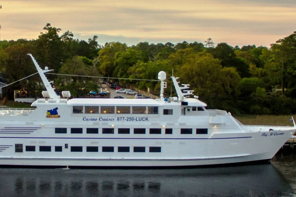 An exterior shot showcasing a casino cruise ship docked on the water, surrounded by lush trees and set against the backdrop of nearby buildings. The scene conveys a blend of excitement and relaxation, hinting at opportunities for dining, entertainment, and socializing on board.