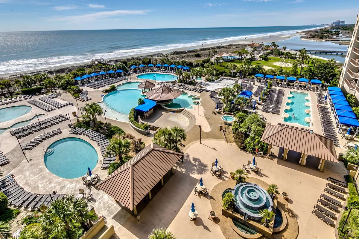 This image captures a sprawling resort pool area featuring multiple swimming pools, sun loungers, palm trees, poolside cabanas, and a breathtaking ocean view. The mood is carefree and vacation-like, inviting relaxation and social gatherings.
