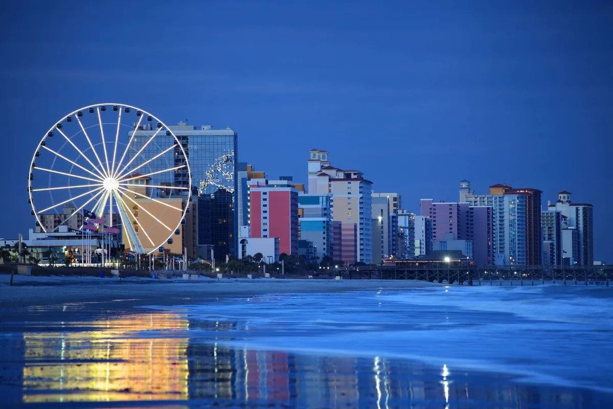 A vibrant exterior scene featuring a ferris wheel, colorful buildings, and ocean waves rolling onto the beach. The festive atmosphere is enhanced by the evening light, hinting at amusement rides, boardwalk excitement, and fun-filled nights by the shore.